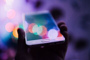 A hand holding a smartphone whose screen reflects blurred colored lights on a purple background